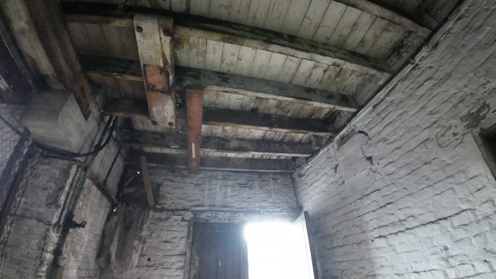 Interior of the old storage and clerical building showing debris, overhead pipework, and structural condition across the three-storey building