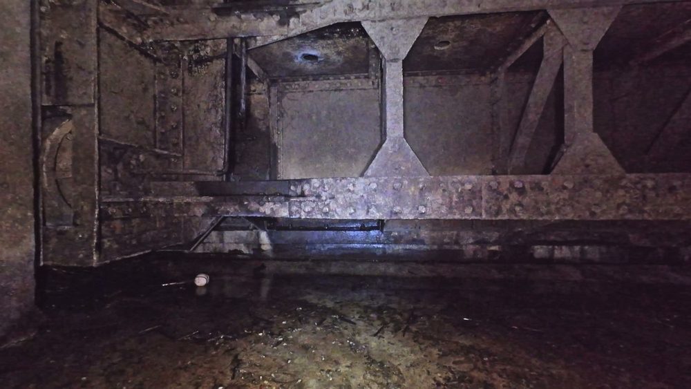 The riveted gate structure and bracing at the top of the drained penstock, lit by the drone