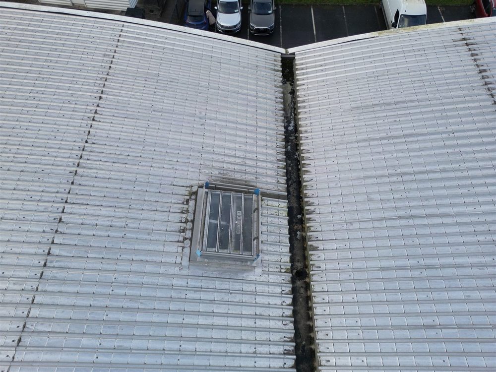 Drone view of adjacent commercial roof sections showing a rooflight, gutter valley, and debris accumulation along the join between two roof pitches