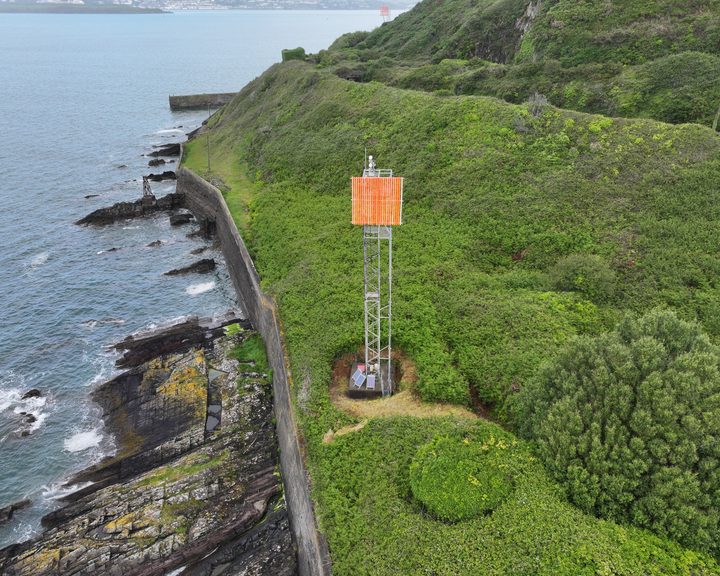 An aerial view of a tall galvanised steel navigation tower carrying an orange board, standing on steep ground above the water at the entrance to Cork Harbour