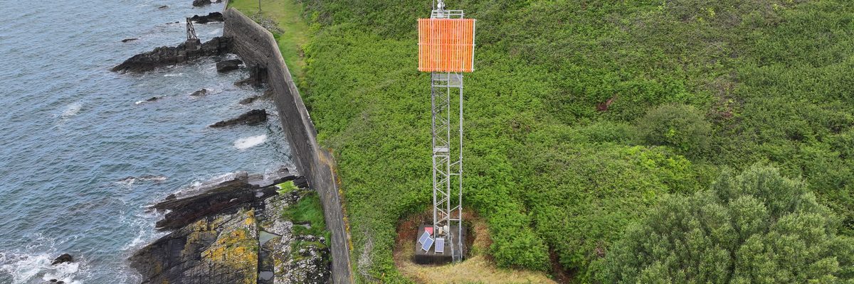 An aerial view of a tall galvanised steel navigation tower carrying an orange board, standing on steep ground above the water at the entrance to Cork Harbour
