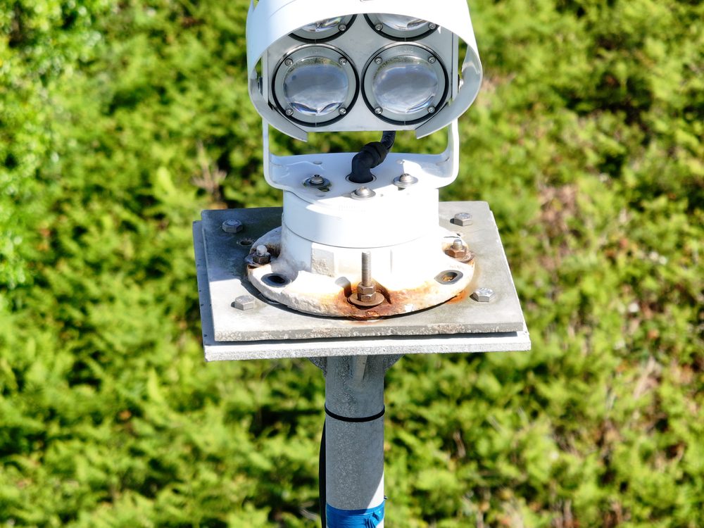 A white multi-lens lighting unit mounted on a grey metal pedestal, with green foliage in the background