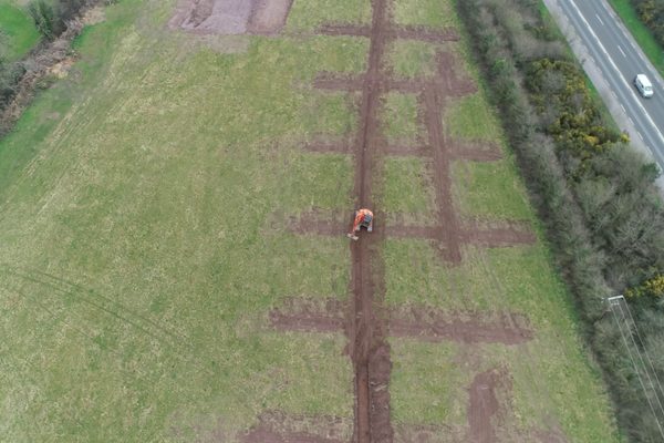 A drone view of an excavator working a pattern of excavated trenches across a green field